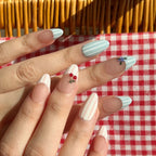 Close-up of a hand with decorative fake nails on a red and white checkered tablecloth.  Pink and white striped & blue and white striped nail designs featuring 3D cherry and blueberry sculpted. LuxeCharm press on nails