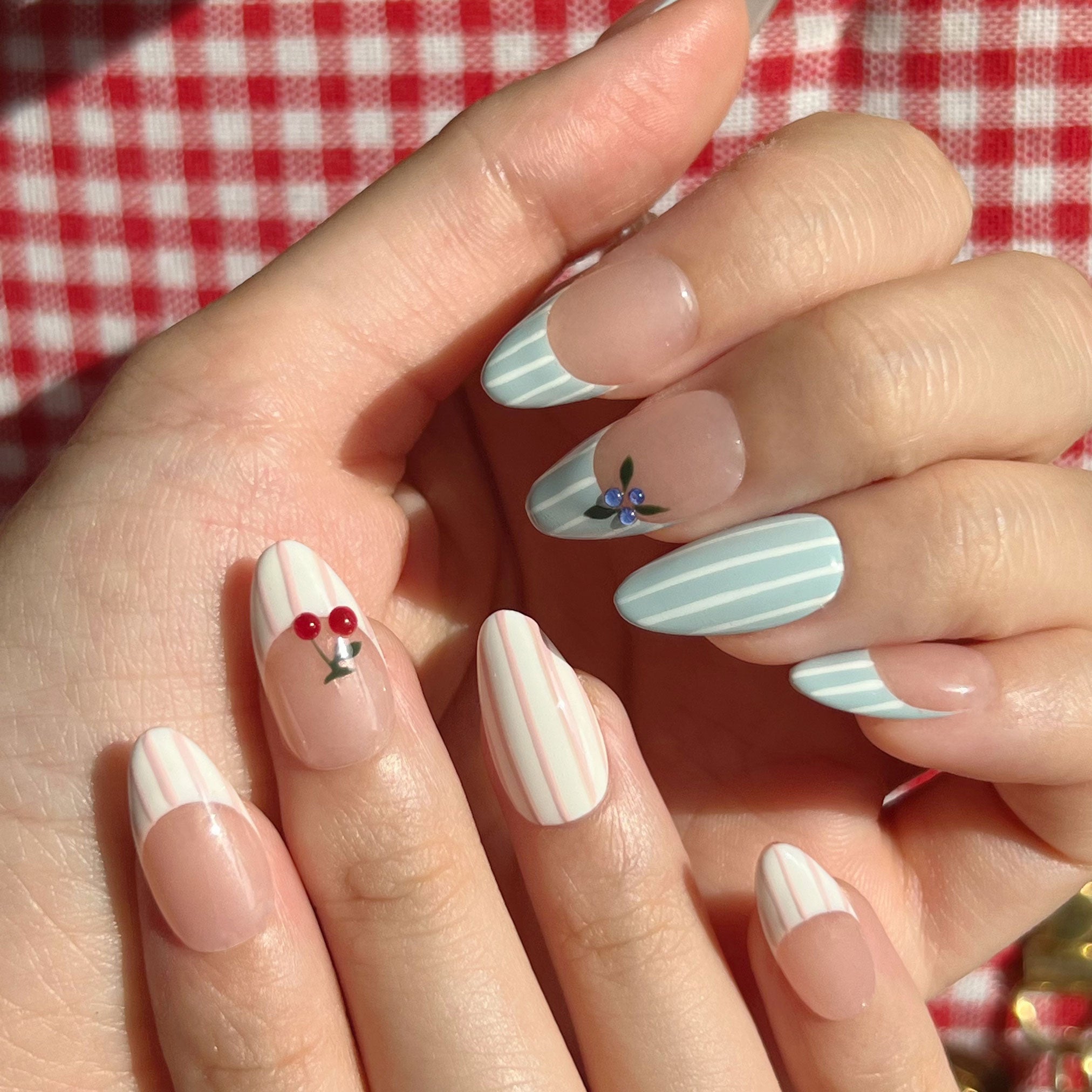 Close-up of a hand with decorative nail art on a red and white checkered tablecloth background.  Pink and white striped & blue and white striped nail designs featuring 3D cherry and blueberry sculpted. LuxeCharm press on nails