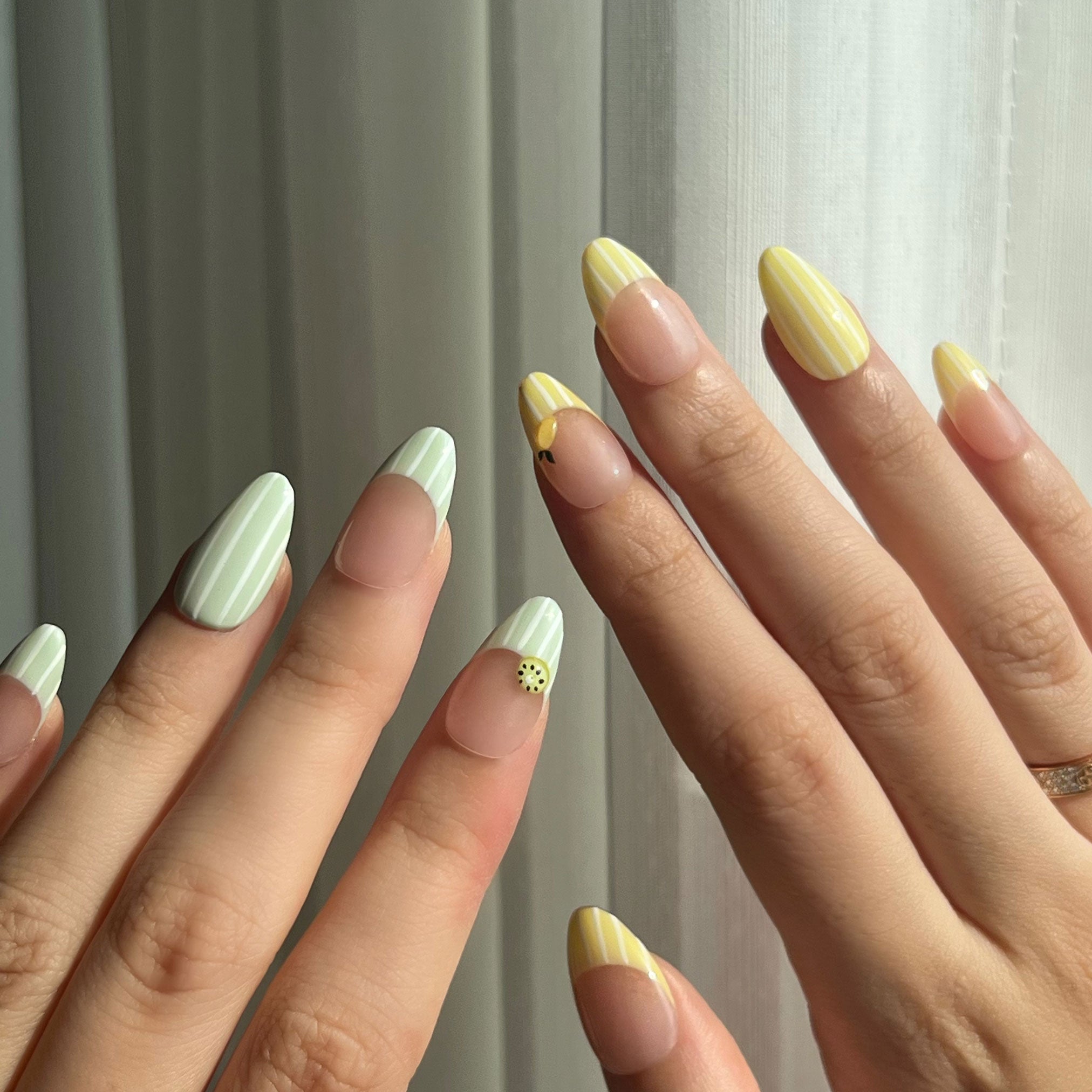 Close-up of hands with green and yellow fake nails against a neutral background. Avocado green and white striped & yellow and white striped nail designs featuring 3D kiwi and lemon sculpted. LuxeCharm press on nails