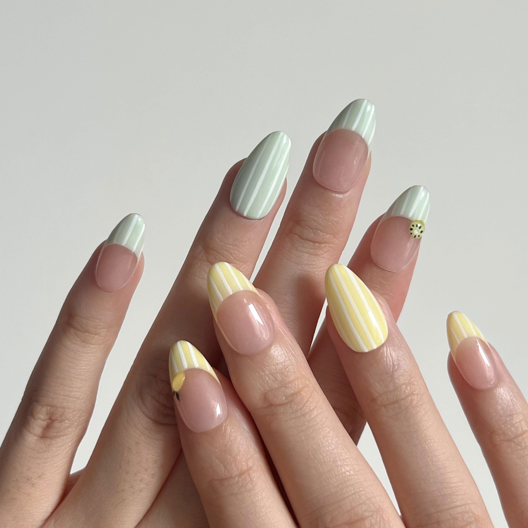 Hand with striped nail art on a plain background. Avocado green and white striped & yellow and white striped nail designs featuring 3D kiwi and lemon sculpted. LuxeCharm press on nails
