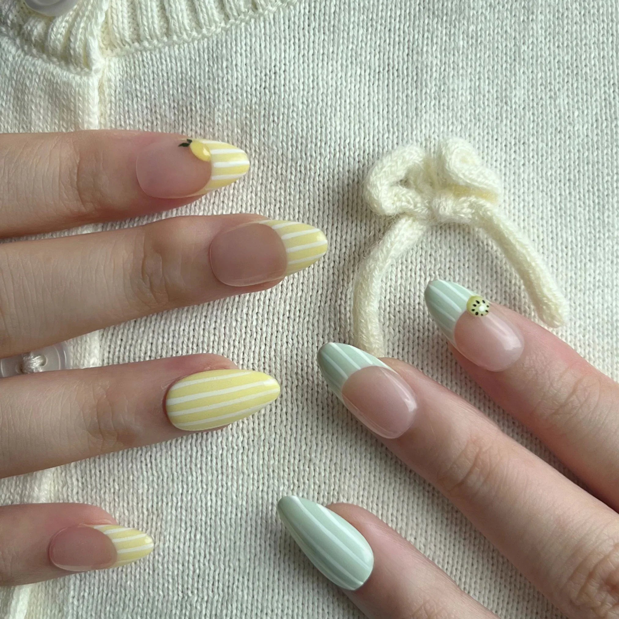 Close-up of hands with pastel nail designs on a textured fabric background. Avocado green and white striped & yellow and white striped nail designs featuring 3D kiwi and lemon sculpted. LuxeCharm press on nails
