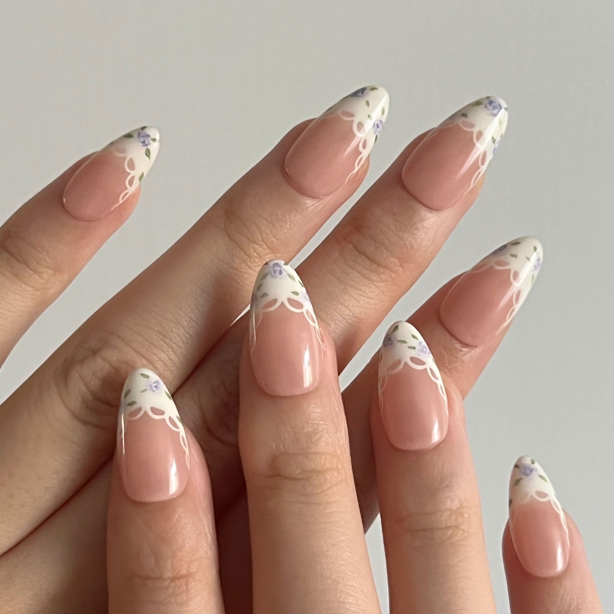 Hand with French tips featuring purple floral designs on a neutral background. Set of purple coquette nail art designs with hand-painted purple floral patterns. LuxeCharm press on nails