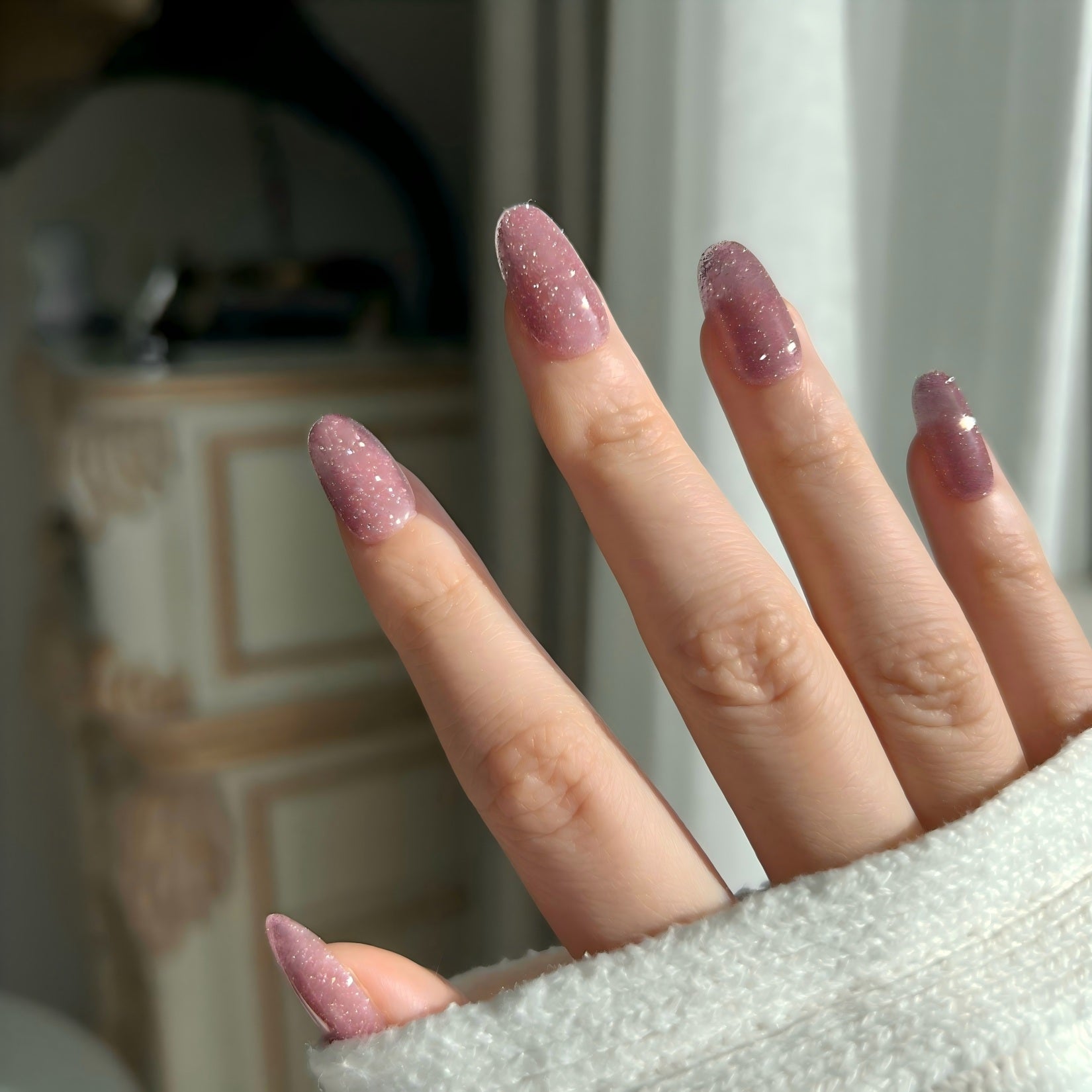 Close-up of a hand with glittery pink nails in a blurred indoor setting. LuxeCharm press on nails