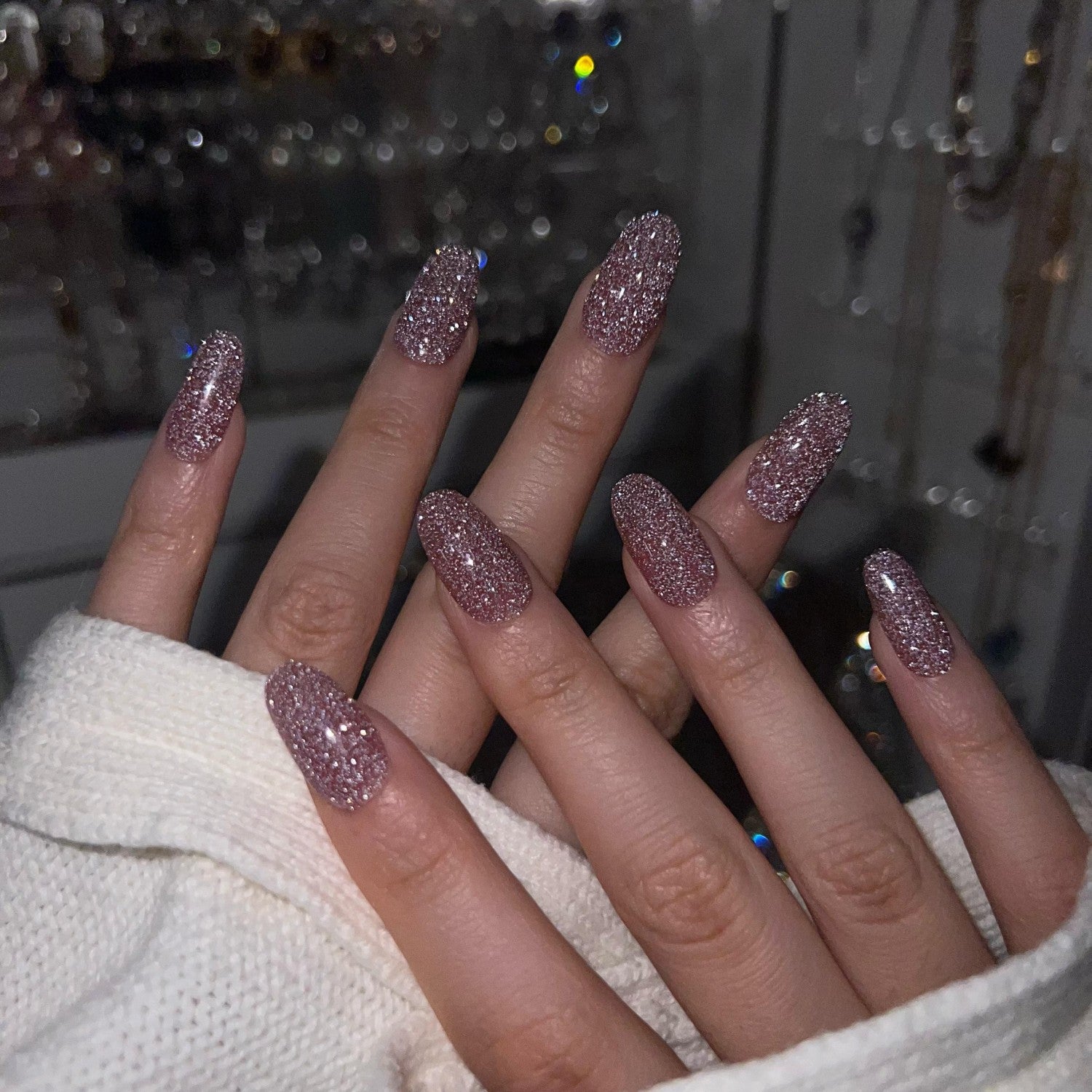 Close-up of hands with glittery pink nails against a blurred background at night. LuxeCharm press on nails 