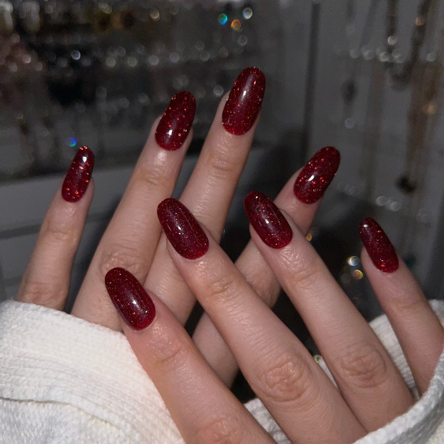 Close-up of a hand with red glitter press-on nails against a blurred background at night. LuxeCharm press on nails