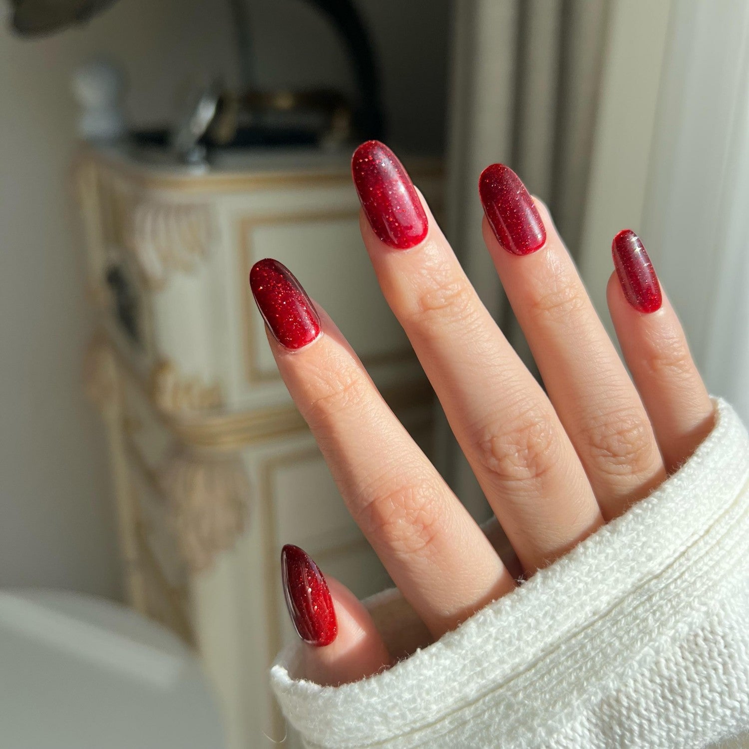 Close-up of a hand with red glitter press on nails against a blurred indoor background