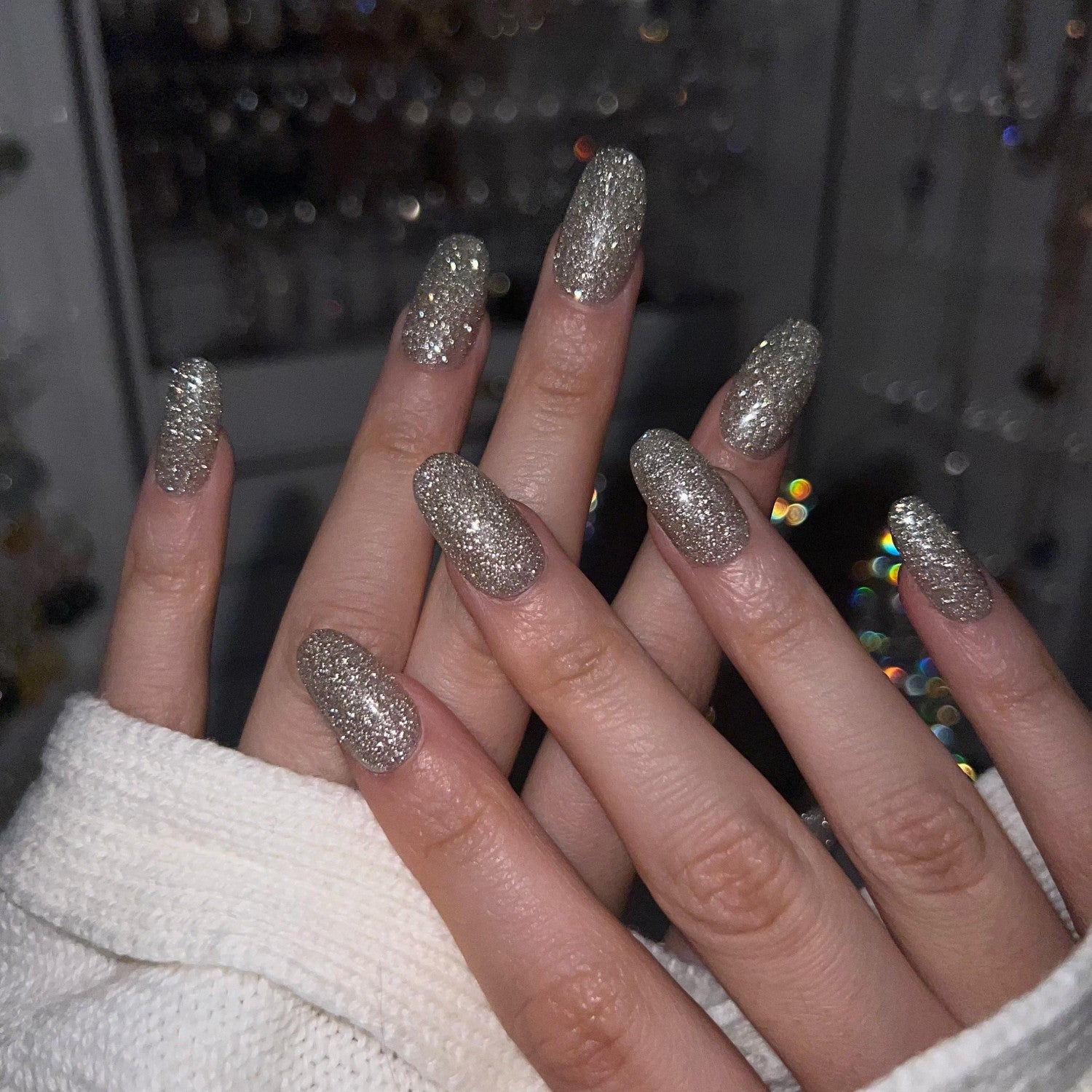 Close-up of hands with silver glitter nails against a blurred background at night.