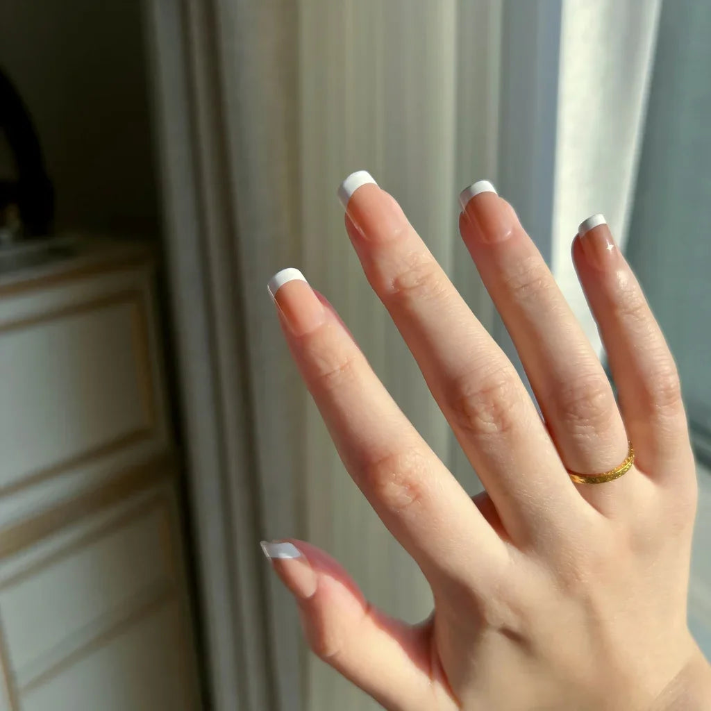 Hand with LuxeTip French extra short square nails in mocha color and a gold ring against a neutral background