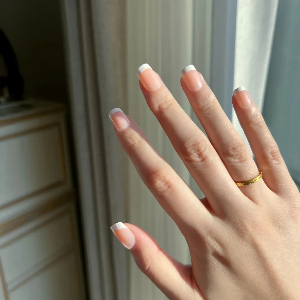 Hand with LuxeTip French extra short square nails in peach color and a gold ring against a neutral background