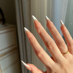 Hand with LuxeTip French long stiletto press on nails in mocha color, wearing a gold ring, against a neutral background.