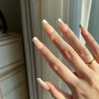 Hand with LuxeTip French medium square press on nails in peach color and a gold ring in front of a neutral background