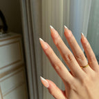 Close-up of a hand with LuxeTip French medium stiletto press on nails in peach color and a gold ring, against a neutral background.