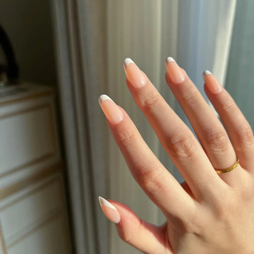 Hand with LuxeTip French short oval nails in peach color and a gold ring against a neutral background