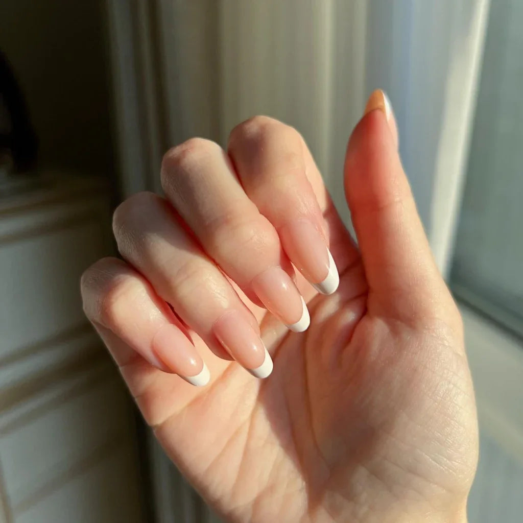 Close-up of a hand with LuxeTip French short oval press on nails with peach color in front of a window.