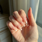 Close-up of a hand with LuxeTip French short oval press on nails with peach color in front of a window.