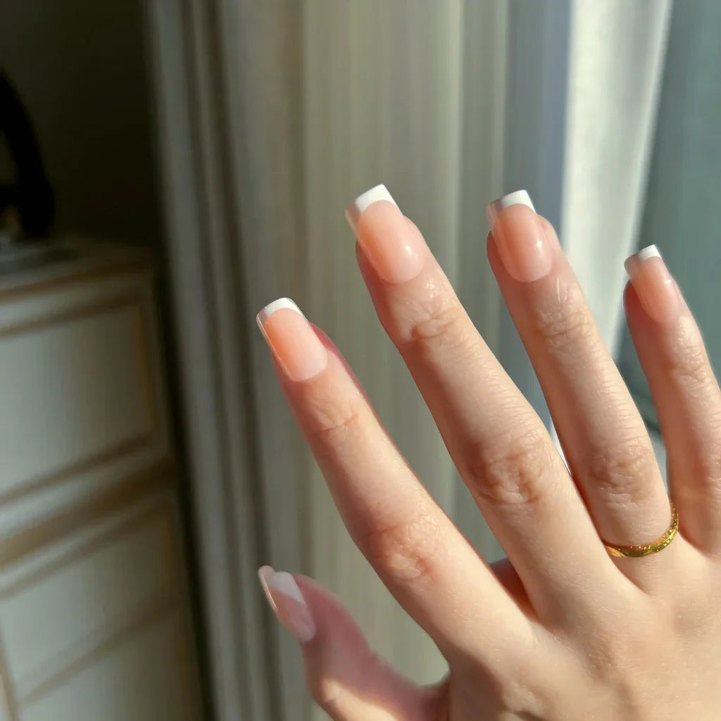 Close-up of a hand with LuxeTip French short square press-on nails in peach color and a gold ring, blurred background